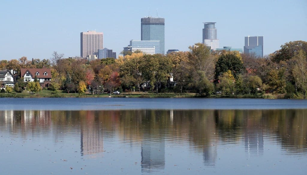Summer view across Lake of the Isles in Minneapolis, with a tree-lined, vegetated shoreline and the downtown skyline reflected in calm water