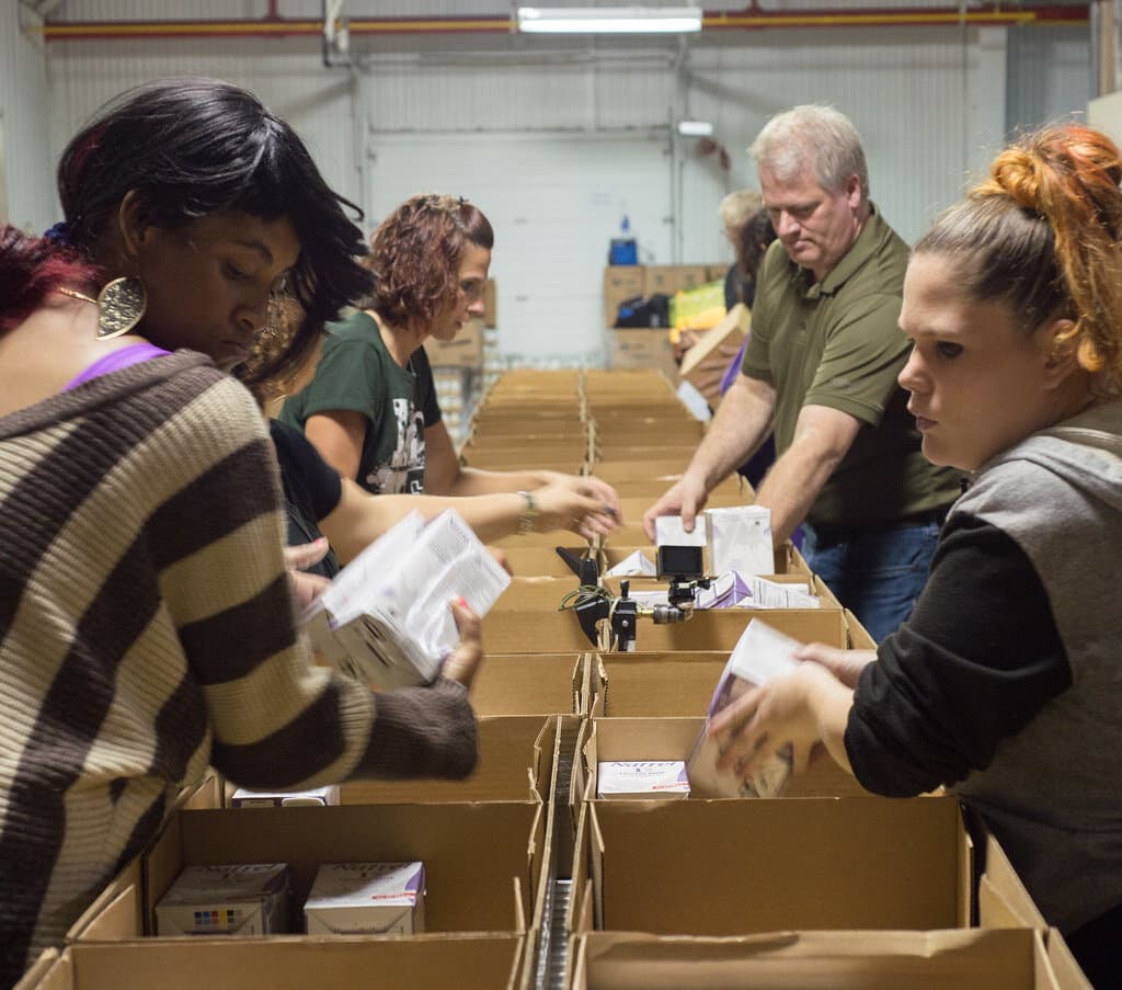 Volunteers in casual clothes pack groceries into cardboard boxes at a community food distribution.